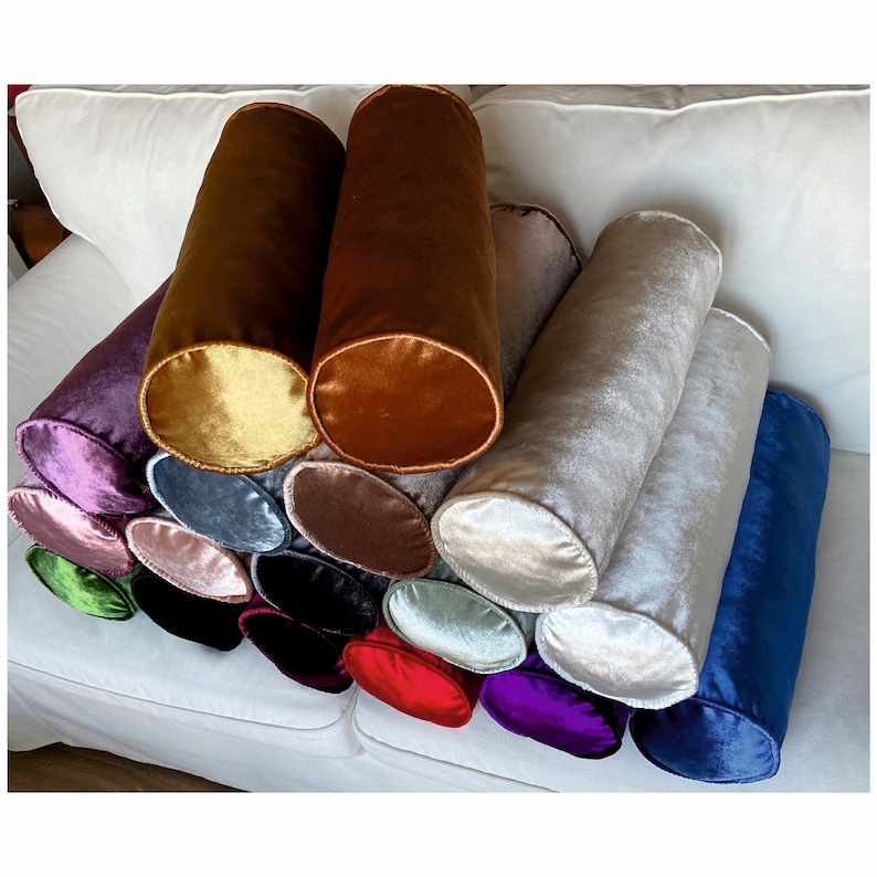 K&ouml;nnte beinhalten: Eine Sammlung von 15 zylindrischen Samt-Kissen in verschiedenen Farben, darunter Gold, Braun, Lila, Blau, Gr&uuml;n und Rosa. Die Kissen sind in einem Halbkreis auf einem wei&szlig;en Sofa angeordnet.
