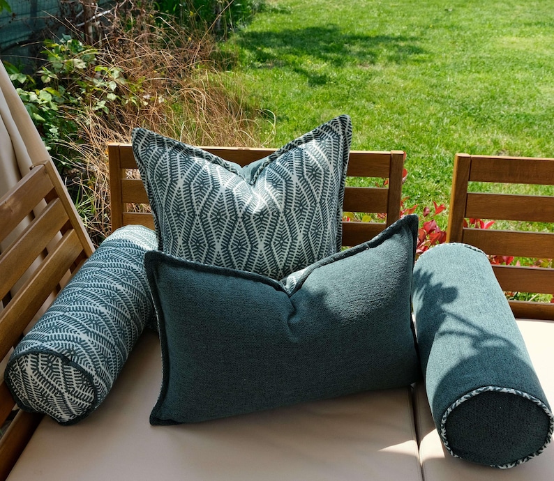 K&ouml;nnte beinhalten: Eine Anordnung von Outdoor-Kissen und -Rollen in T&uuml;rkis und Wei&szlig;. Die Kissen weisen geometrische Muster und unifarbene Texturen mit dunklen Paspeln auf. Die Anordnung befindet sich auf einer hellbeigen Oberfl&auml;che, mit einem Holzstuhl im Hintergrund.