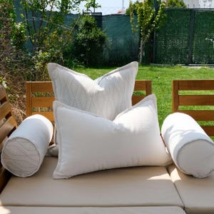K&ouml;nnte beinhalten: Eine Ansammlung wei&szlig;er und hellgrauer Zierkissen und -rollen, die auf einem beigen Outdoor-Sofa angeordnet sind. Die Kissen haben verschiedene Texturen, darunter ein gemustertes Design auf einigen. Die Umgebung ist ein sonniger Au&szlig;enbereich mit Gr&uuml;n.