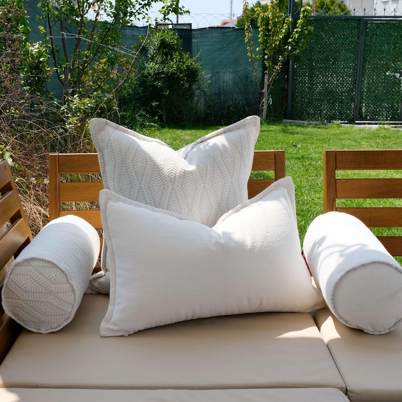 K&ouml;nnte beinhalten: Eine Ansammlung wei&szlig;er und hellgrauer Zierkissen und -rollen, die auf einem beigen Outdoor-Sofa angeordnet sind. Die Kissen haben verschiedene Texturen, darunter ein gemustertes Design auf einigen. Die Umgebung ist ein sonniger Au&szlig;enbereich mit Gr&uuml;n.