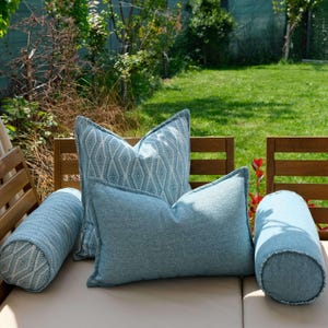 K&ouml;nnte beinhalten: Eine Auswahl an dekorativen Kissen in Blau- und Wei&szlig;t&ouml;nen. Das Set umfasst zwei quadratische Kissen mit gemusterten und unifarbenen Designs sowie zwei zylindrische Nackenrollen. Die Kissen sind auf einer beigen Oberfl&auml;che angeordnet, mit gr&uuml;nem Hintergrund.