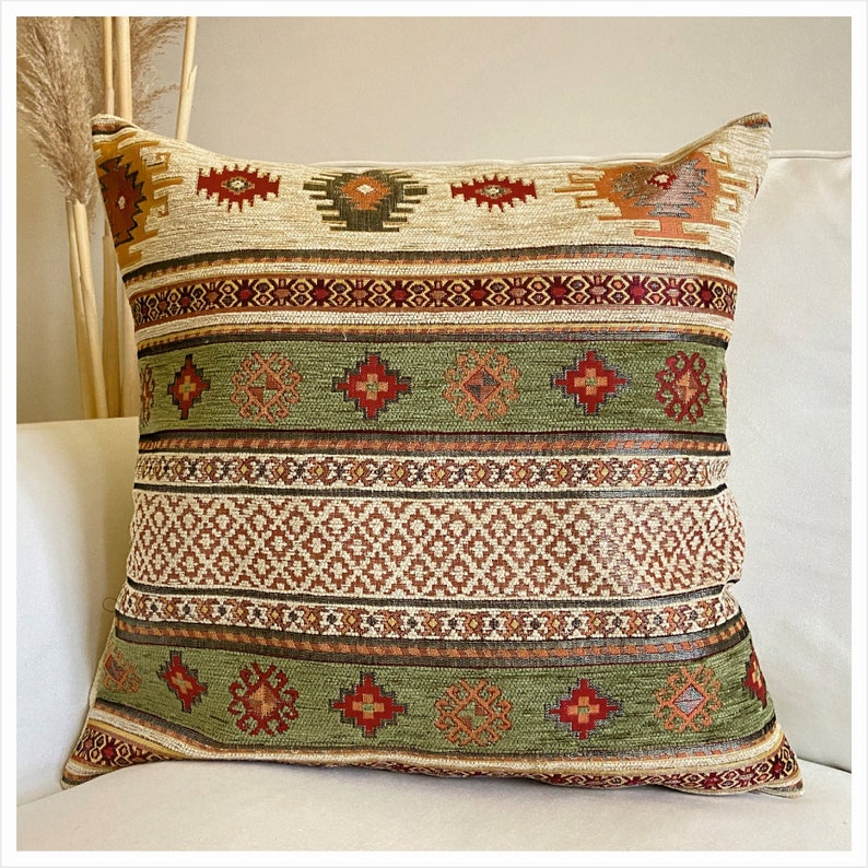 Puede incluir: Coj&iacute;n decorativo cuadrado con un dise&ntilde;o tejido. El coj&iacute;n presenta una parte superior e inferior beige con patrones geom&eacute;tricos en rojo, naranja y marr&oacute;n. La secci&oacute;n central es verde con patrones similares. El coj&iacute;n est&aacute; sobre una superficie blanca.