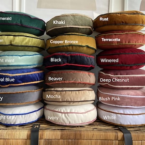 May include: A stack of 15 round velvet cushions in various colours, including emerald green, khaki, rust, green, terracotta, copper bronze, light mint, black, winered, royal blue, beaver, deep chestnut, grey, mocha, blush pink, light grey, ivory, and white.