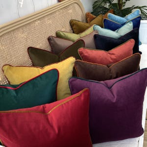 May include: A collection of velvet throw pillows with contrasting trim in various colors, including mustard yellow, forest green, burgundy, and purple. The pillows are arranged in a row on a wicker chair.