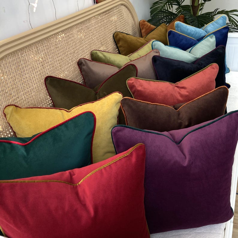 May include: A collection of velvet throw pillows with contrasting trim in various colors, including mustard yellow, forest green, burgundy, and purple. The pillows are arranged in a row on a wicker chair.