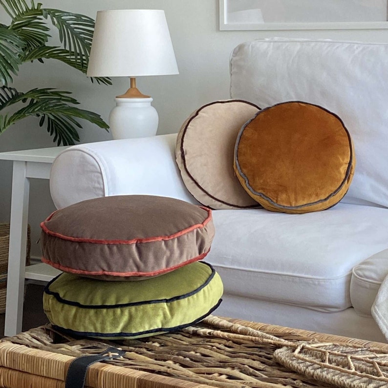 Pu&ograve; includere: Tre cuscini rotondi in velluto con passamaneria a contrasto. I cuscini sono in tonalit&agrave; di marrone, beige e verde. I cuscini sono su un divano bianco e un tavolino in vimini.