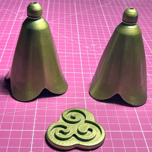 May include: Two gold-colored, bell-shaped, decorative finials with a three-leaf design at the bottom. A gold-colored, three-leaf design is in the foreground.