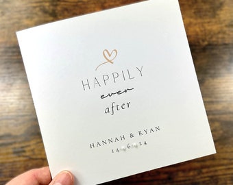 May include: White card with the text "Happily ever after" in black script font. A gold heart symbol is above the text. Below the text is "HANNAH & RYAN" in black font and "14.06.24" in black font with small white pearl-like dots between the numbers.