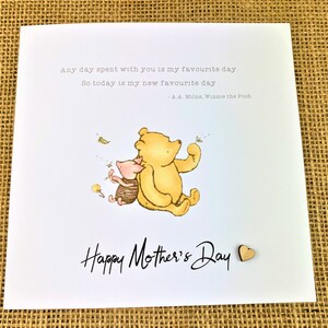 May include: A white greeting card with the text "Happy Mother's Day" in black script. The card features a watercolor illustration of Winnie the Pooh and Piglet. A small wooden heart is attached to the card.