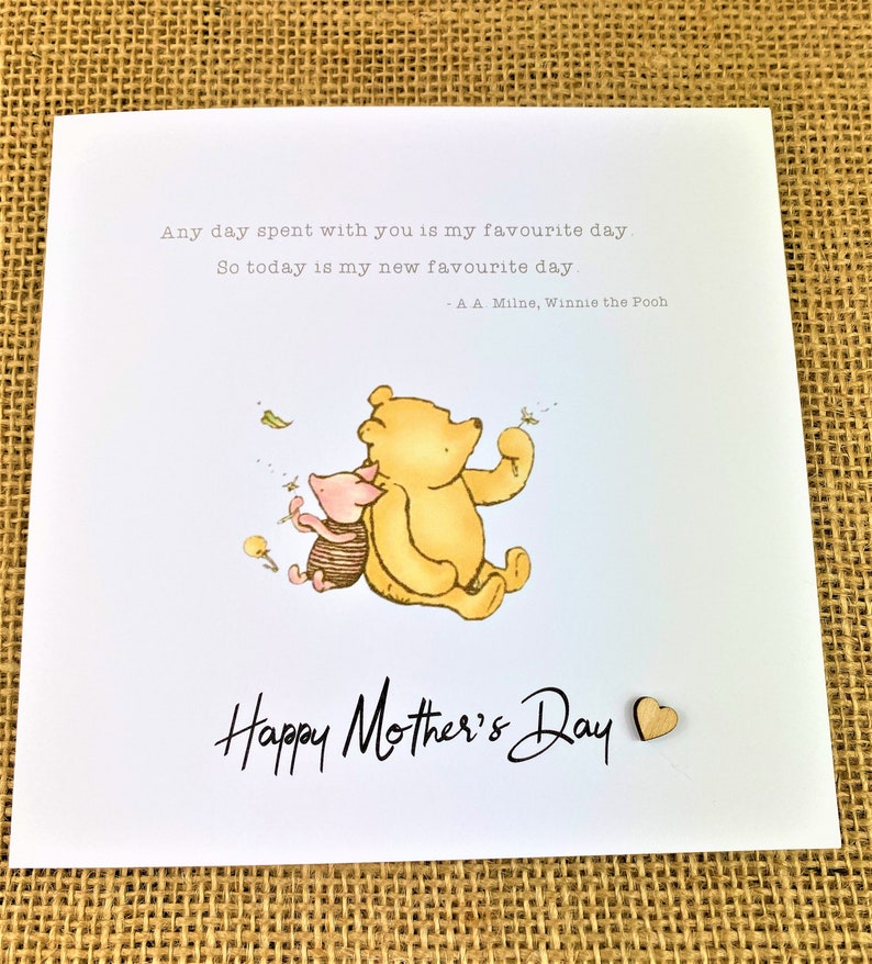 May include: A white greeting card with the text "Happy Mother's Day" in black script. The card features a watercolor illustration of Winnie the Pooh and Piglet. A small wooden heart is attached to the card.