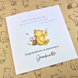 May include: A white card with a Winnie the Pooh and Piglet illustration. The card has the text "Sometimes the smallest things take up the most room in your heart - A.A. Milne, Winnie the Pooh" and "Congratulations on becoming a Grandma".