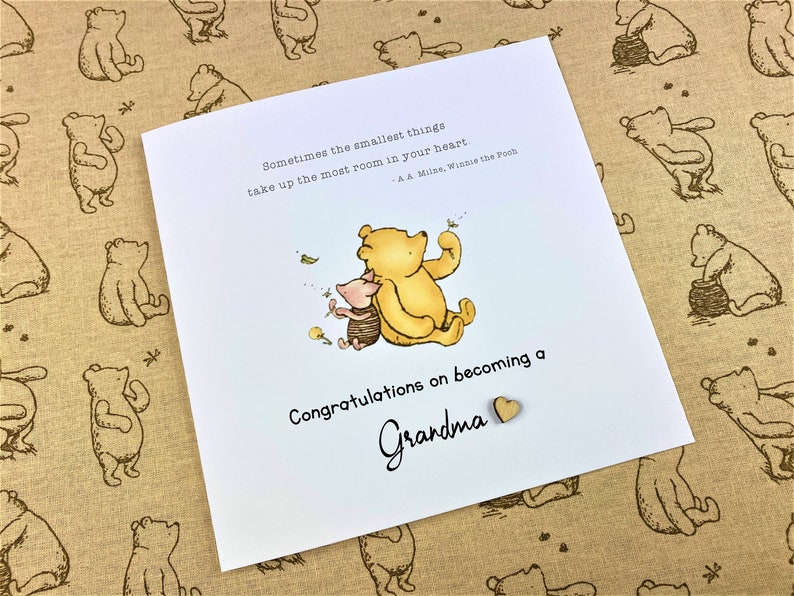May include: A white card with a Winnie the Pooh and Piglet illustration. The card has the text "Sometimes the smallest things take up the most room in your heart - A.A. Milne, Winnie the Pooh" and "Congratulations on becoming a Grandma".