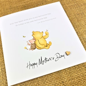 May include: A white greeting card with the text "Happy Mother's Day" in black script. The card features an illustration of Winnie the Pooh and Piglet. A small wooden heart is attached to the card.