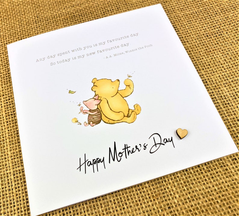 May include: A white greeting card with the text "Happy Mother's Day" in black script. The card features an illustration of Winnie the Pooh and Piglet. A small wooden heart is attached to the card.