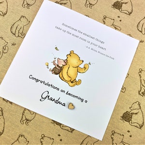 May include: A white card with a Winnie the Pooh and Piglet illustration. The card has a quote from A.A. Milne, Winnie the Pooh, "Sometimes the smallest things take up the most room in your heart." The card says "Congratulations on becoming a Grandma" with a small wooden heart.