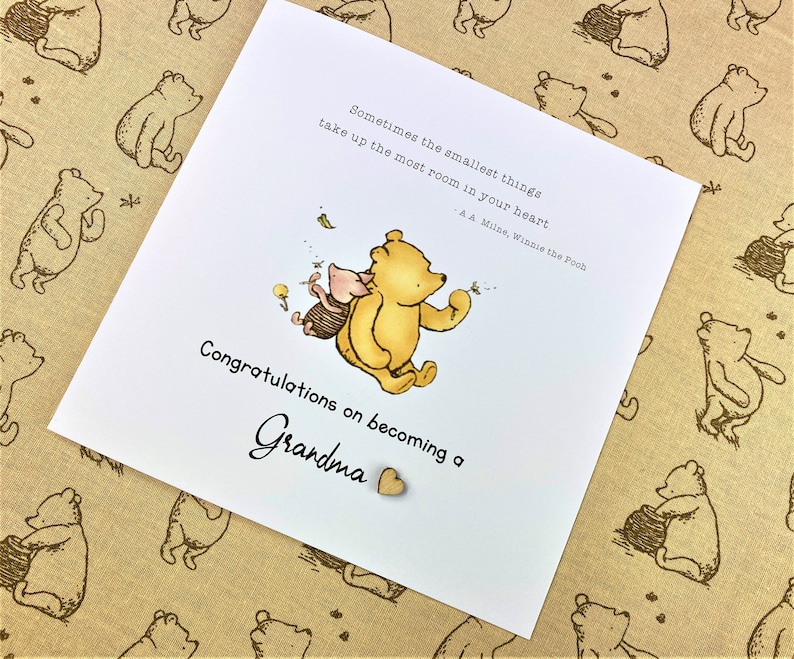 May include: A white card with a Winnie the Pooh and Piglet illustration. The card has a quote from A.A. Milne, Winnie the Pooh, "Sometimes the smallest things take up the most room in your heart." The card says "Congratulations on becoming a Grandma" with a small wooden heart.