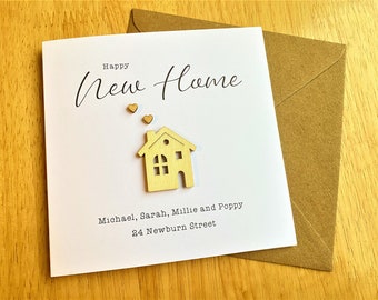 May include: A white card with the text "Happy New Home" in black cursive writing. There are three small gold hearts above the text and a small wooden house cutout below the text. The card also includes the text "Michael, Sarah, Millie and Poppy 34 Newburn Street" in black.