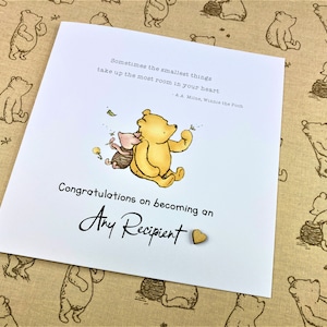 May include: A white card with a Winnie the Pooh and Piglet illustration. The card has the text "Sometimes the smallest things take up the most room in your heart - A A Milne, Winnie the Pooh" and "Congratulations on becoming an Any Recipient".