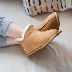 May include: A pair of brown suede slippers with white fur lining. The slippers are being worn by a person sitting on a white fluffy surface.