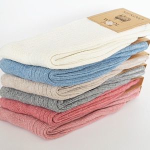 Könnte beinhalten: Ein Stapel von sechs Paar gerippten Merinowollsocken in verschiedenen Farben: Weiß, Hellblau, Beige, Grau und zwei Rosatönen. Jedes Paar ist gefaltet und hat ein braunes Papieretikett mit der Aufschrift "MERINO WOOL".