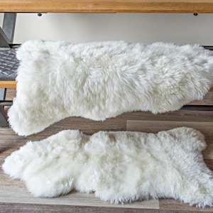 May include: Two white sheepskin rugs, one on top of the other, on a wooden floor. The top rug is draped over a wooden bench with a black metal frame.