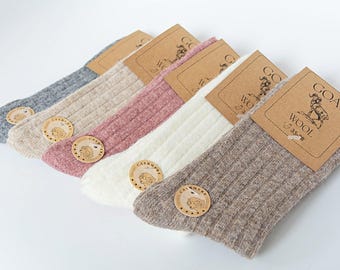 May include: Five pairs of ribbed wool socks in shades of grey, beige, pink, white, and brown. Each pair has a wooden tag with a logo and the words "GOAT WOOL". The socks are displayed on a white surface.