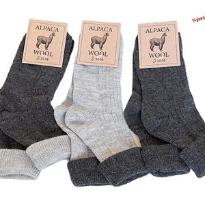 Può includere: Tre paia di calzini in lana di alpaca in diverse tonalità di grigio, con polsini grigio chiaro a contrasto. Ogni paio ha un'etichetta con un'illustrazione di alpaca e le parole "ALPACA WOOL". Il testo "Spring/Summer Collection!" è visibile.