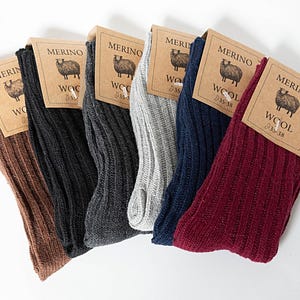 Può includere: Sei paia di calzini in lana merino a coste in vari colori: marrone, nero, grigio scuro, grigio chiaro, blu navy e bordeaux. Ogni paio ha un'etichetta con un'illustrazione di pecora e la scritta "MERINO WOOL".
