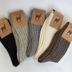 May include: Five pairs of alpaca wool socks in different colors: black, gray, white, brown, and beige. Each sock has a brown tag with the words "Alpaca Wool" and a small image of an alpaca.