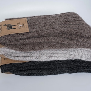 Puede incluir: Tres pares de calcetines de lana de alpaca en marrón, gris y negro. Los calcetines están doblados y apilados uno encima del otro. El par de calcetines superior tiene una etiqueta de papel marrón con el texto "Alpaca Wool" y una imagen de una alpaca.
