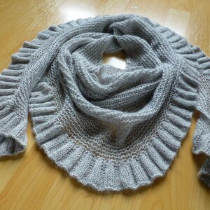 Könnte beinhalten: Ein grauer gestrickter Schal mit einem gekräuselten Rand. Der Schal ist aus einem weichen, leichten Garn gefertigt und hat ein zartes, luftiges Gefühl.