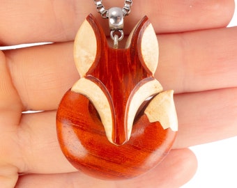 May include: A wooden fox pendant necklace with a silver chain. The fox is carved from two types of wood, one light and one dark, creating a contrasting pattern. The pendant is a detailed representation of a fox with a curled tail.
