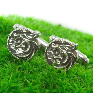 Può includere: Un paio di gemelli d'argento con un design a testa di lupo. I gemelli sono su una superficie verde ricoperta di muschio.