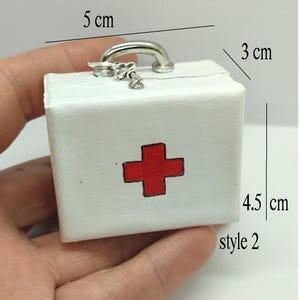 以下が含まれることがあります： 前面に赤い十字架が描かれた白い救急箱。箱には銀色の取っ手と小さなチェーンが付いています。箱のサイズは幅5cm、高さ4.5cm、奥行き3cmです。