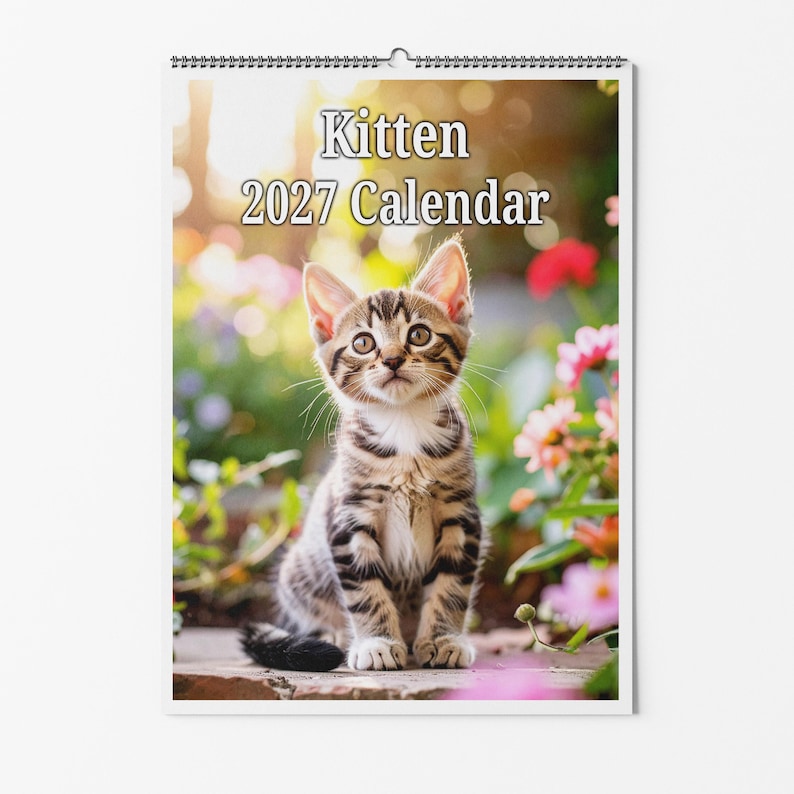 以下が含まれることがあります： Kitten 2027 Calendarというタイトルの壁掛けカレンダー。屋外に座る茶色のトラ猫の子猫の写真が特徴です。子猫には濃い縞模様、大きな目、尖った耳があります。背景には、日光を浴びたぼやけた色とりどりの花と緑が写っています。