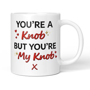 May include: White ceramic mug with the text "YOU'RE A Knob BUT YOU'RE My Knob:" in black and red lettering. The design includes small red hearts and starburst accents. The mug has a white handle.