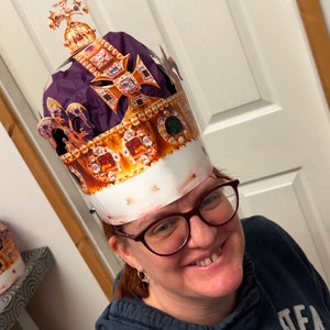 May include: A person wearing a gold and purple paper crown with jewels and a white background. The crown is shaped like a traditional royal crown.