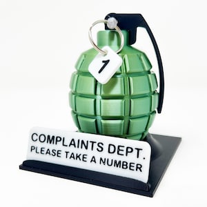 May include: A green grenade-shaped desk ornament with a black handle and base. A white tag with the number "1" hangs from a ring on top. The base has a sign that reads "COMPLAINTS DEPT. PLEASE TAKE A NUMBER."