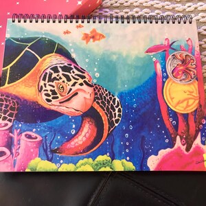 May include: Spiral-bound notebook featuring a vibrant underwater scene. A detailed illustration of a sea turtle, coral, and fish in shades of blue, orange, and pink. The artwork has a whimsical, artistic style, perfect for note-taking or sketching.