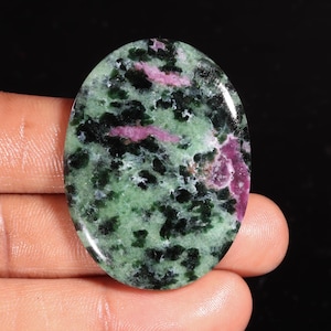 Puede incluir: Un cabujón de piedra preciosa pulida de forma ovalada con un patrón moteado de minerales verde claro, verde oscuro y negro con parches distintos de inclusiones de color rosa magenta vibrante en toda la superficie lisa y reflectante.