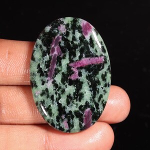 Puede incluir: Una piedra preciosa pulida de forma ovalada con un patrón moteado de inclusiones minerales en negro profundo y verde claro, acentuada por varias formaciones cristalinas de color magenta, distintas y alargadas, sobre la superficie lisa.