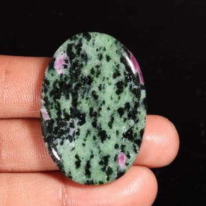 Puede incluir: Una piedra preciosa pulida de forma ovalada con un patrón moteado de inclusiones minerales de color verde claro y verde oscuro, acentuado por pequeñas manchas distintas de color rosa magenta vibrante en la superficie lisa y brillante.
