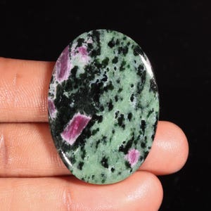 Op de afbeelding: Een ovaalvormige gepolijste edelsteen met een gevlekt patroon van lichtgroene, donkergroene en zwarte mineraalinklusies, geaccentueerd door verschillende opvallende, levendige magenta-roze kristallijne vlekken verspreid over het gladde oppervlak.