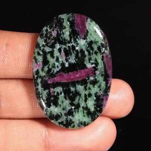 May include: An oval-shaped polished gemstone featuring a mottled pattern of deep black, vibrant green, and magenta-pink inclusions. The surface is smooth and reflective, showcasing a distinct contrast between the dark and light mineral deposits.