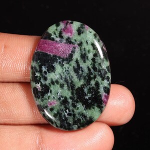 Puede incluir: Una piedra preciosa pulida de forma ovalada con un patrón moteado de verde bosque profundo, negro e inclusiones de color rosa magenta vibrante. La superficie es lisa y reflectante, mostrando una composición mineral natural.