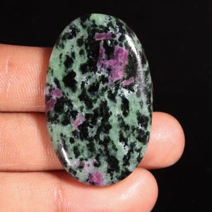 May include: An oval-shaped polished gemstone featuring a mottled pattern of deep forest green, black, and vibrant magenta-pink inclusions. The surface is smooth and reflective, showcasing a natural mineral composition with distinct colour variations.