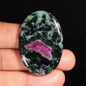 Puede incluir: Una piedra preciosa pulida de forma ovalada que presenta una matriz moteada en verde oscuro y negro con una prominente inclusión irregular de color rosa magenta centrada en la parte inferior. La piedra tiene una superficie lisa y brillante.