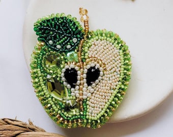 May include: A green and white beaded apple brooch with a green leaf. The brooch is on a white plate.