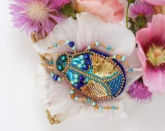 May include: A beaded scarab beetle brooch in shades of blue and gold, adorned with sequins and beads, rests on a white flower petal. The brooch is surrounded by pink and purple flowers and a gold-colored ornate frame.