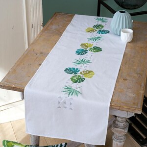 Könnte beinhalten: Ein weißer Tischläufer mit einem gestickten Muster aus grünen Blättern. Der Läufer liegt auf einem Holztisch mit einer Vase und einer kleinen weißen Schale auf dem Tisch.